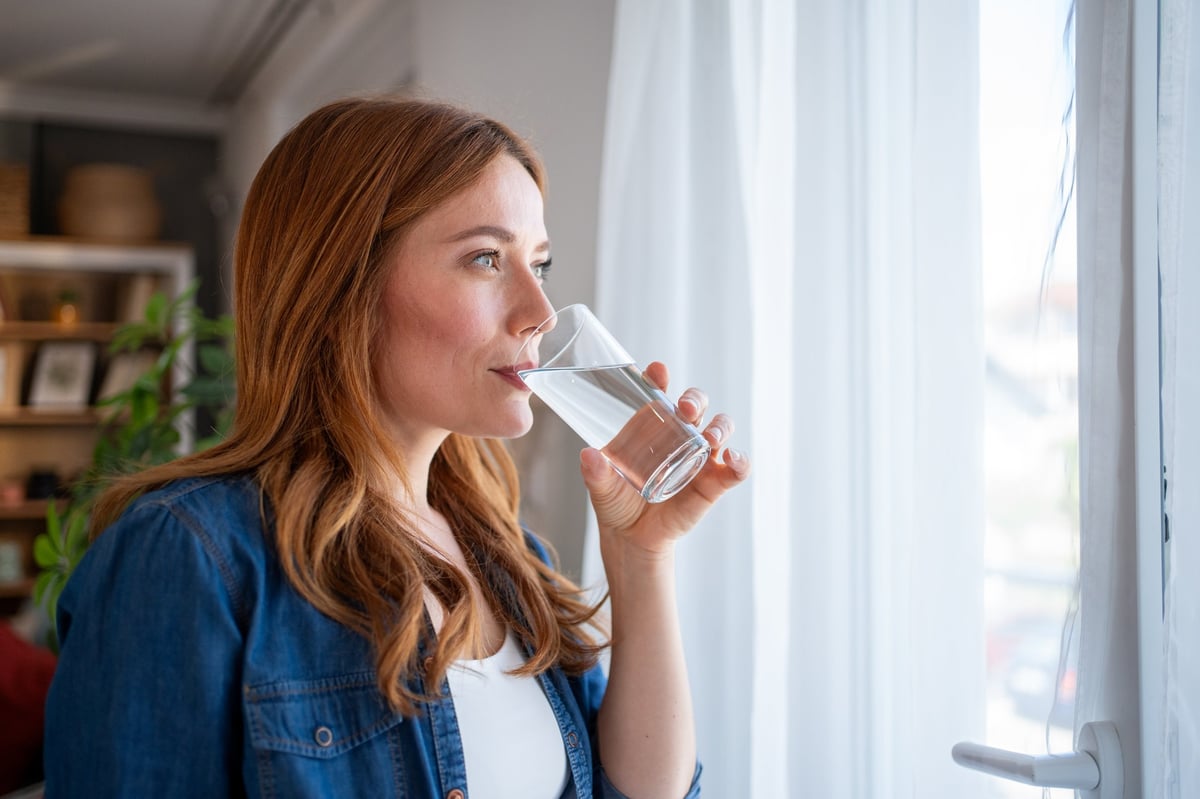 Mid adult woman enjoying a glass of fresh water while standing by the window at home, gazing outside and savoring a peaceful moment of relaxation and hydration