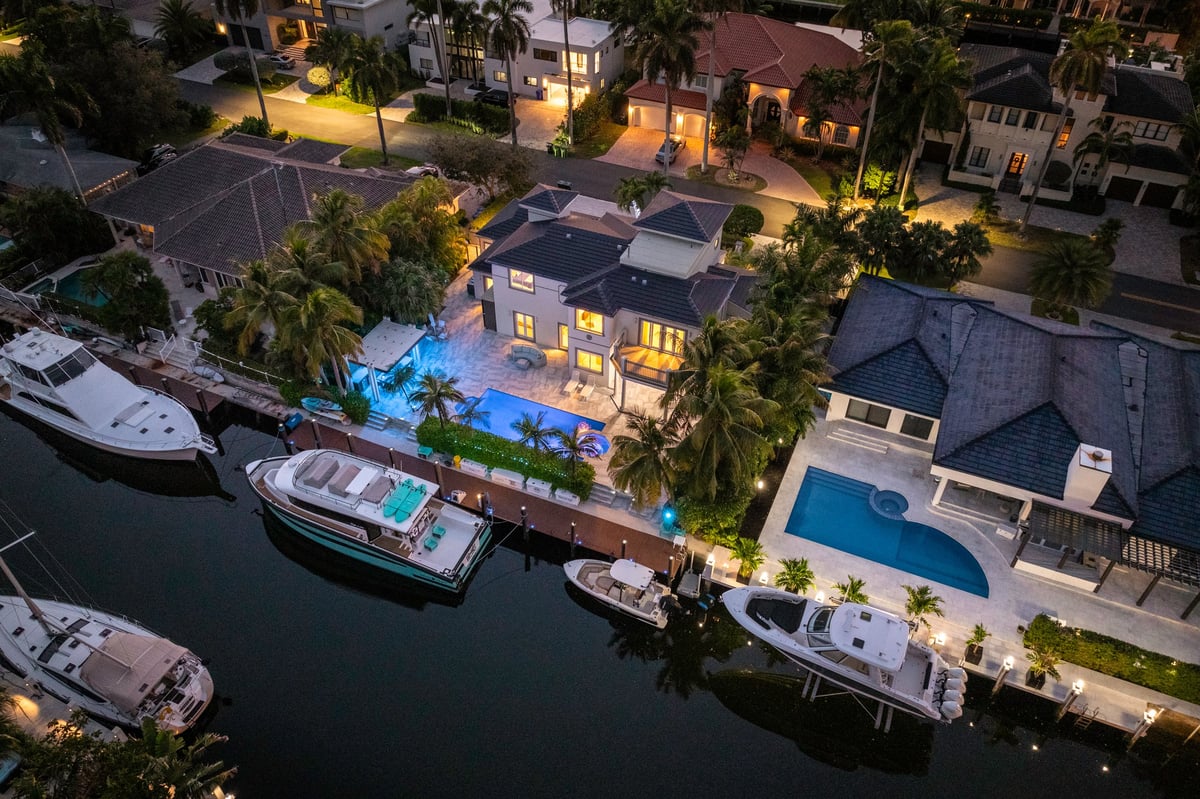Night aerial view of a luxury seaside mansion with private dock and large yachts. Illuminated swimming pool surrounded by palm trees in a tropical setting.