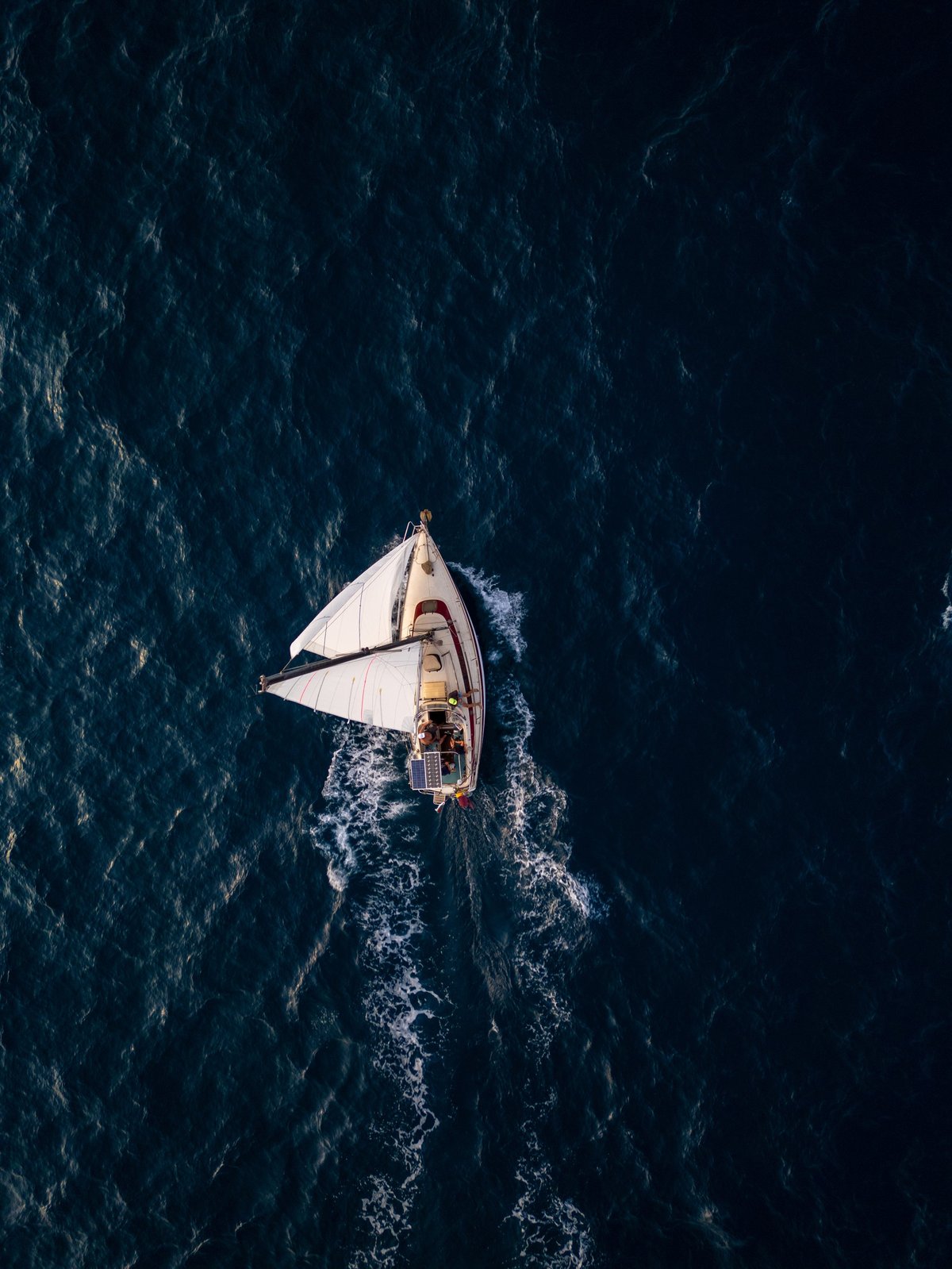 Aerial video of a white sailboat sailing alone on deep blue sea. Calm waves shimmer under sunlight.