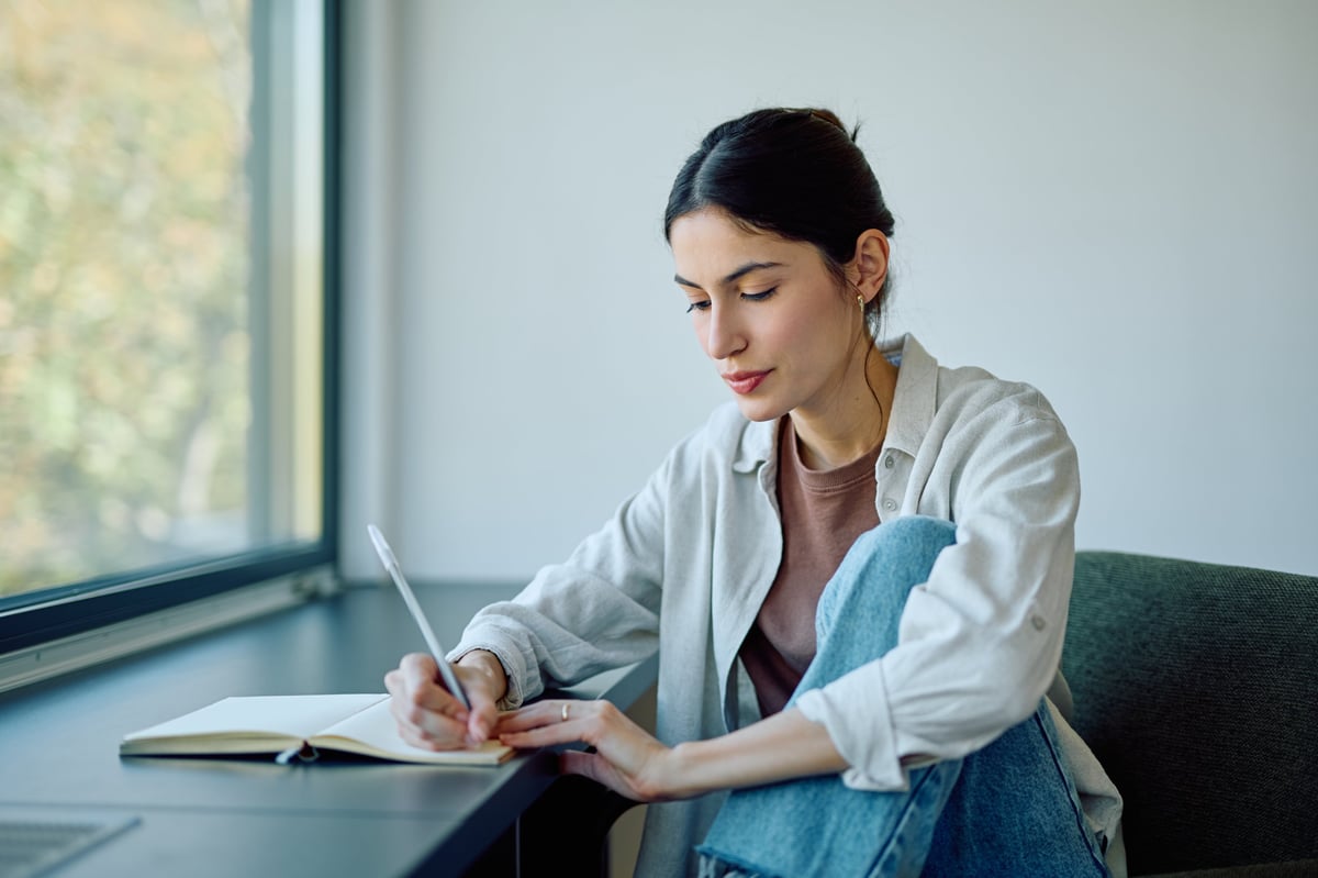 Young woman focusing on writing notes in open notebook, studying or journaling by a window in natural light