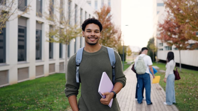 Student with backpack smiling, holding a folder on university campus. Group of students in background, buildings and trees