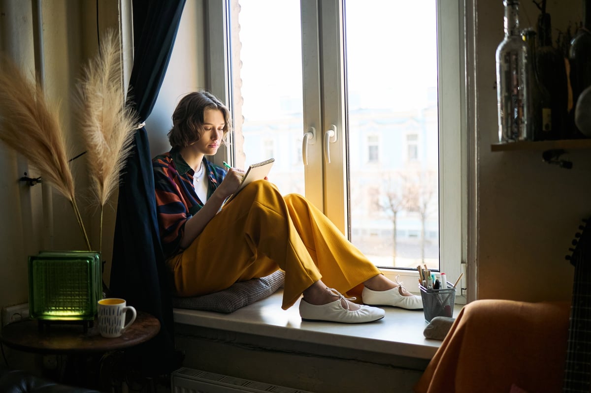Woman sitting on a windowsill writing in a notebook and reflecting