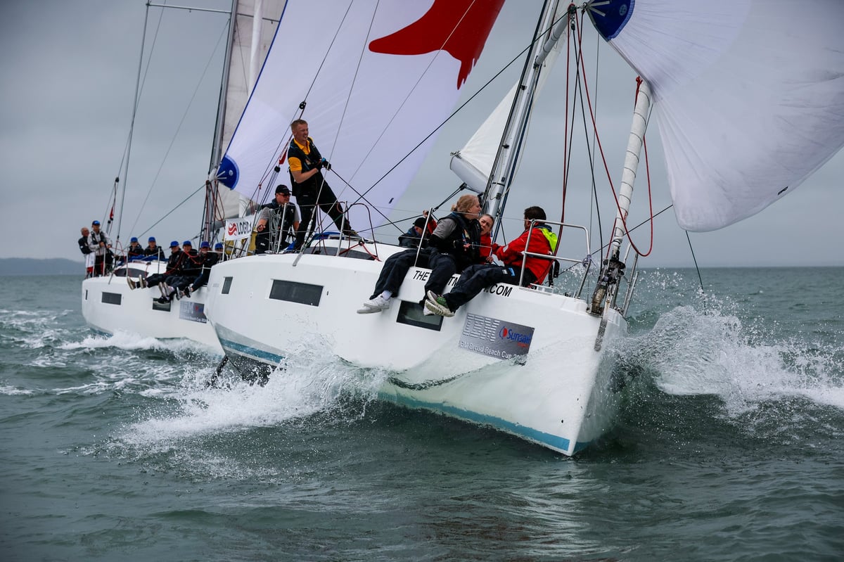 White sailing catamaran with crew members navigating through ocean waves, red and blue flags visible on mast