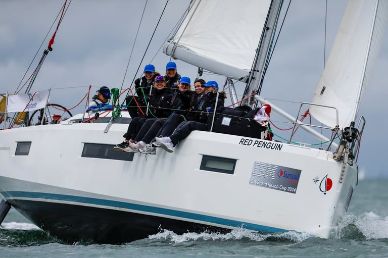 White sailboat with crew members wearing blue jackets sailing through choppy water with white sails raised